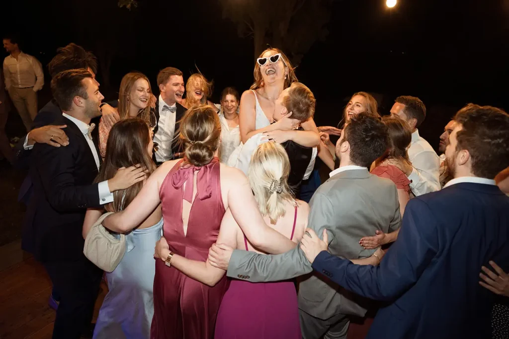 Wedding at I Terzi d'Ombrone. A group of people dressed in formal attire stand in a circle with arms around each other, some laughing and smiling, as one woman in sunglasses is lifted in the center.
