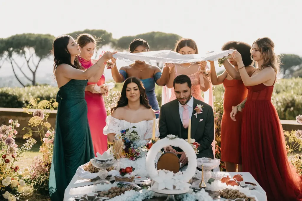 Wedding at Villa Grazioli: A bride and groom sit at a decorated table outdoors while women hold a white cloth above their heads during a traditional wedding ceremony.
