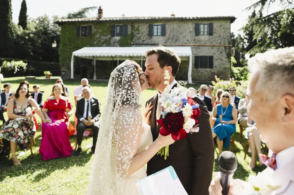 Destination Wedding in Tuscany - A bride and groom kiss during an outdoor wedding ceremony at Podere La Specola, with guests seated behind them and a house in the background.