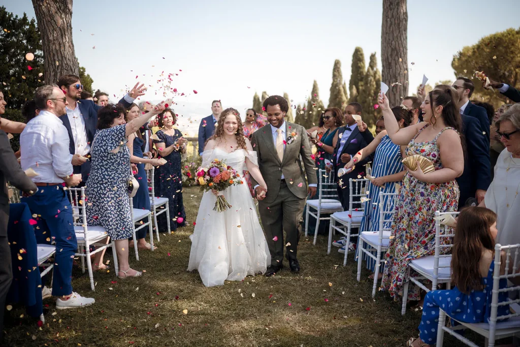 ethiopian wedding tuscany. A bride and groom walk down an outdoor aisle, smiling and holding hands, as guests throw flower petals in celebration.