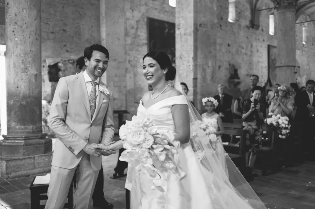 A bride and groom stand together smiling inside the Cathedral of San Cerbone, holding hands and flowers, with guests seated in the background—a perfect setting for a luxury Tuscan wedding.