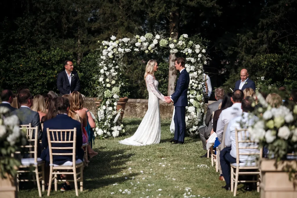 A bride and groom stand under a floral arch exchanging vows at an elegant white green wedding decor Tuscany outdoor wedding venue, with guests seated on either side and officiant nearby.