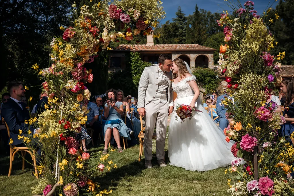 Monte Amiata wedding. A bride and groom kiss under a floral arch at an outdoor wedding ceremony, surrounded by guests seated on the lawn.