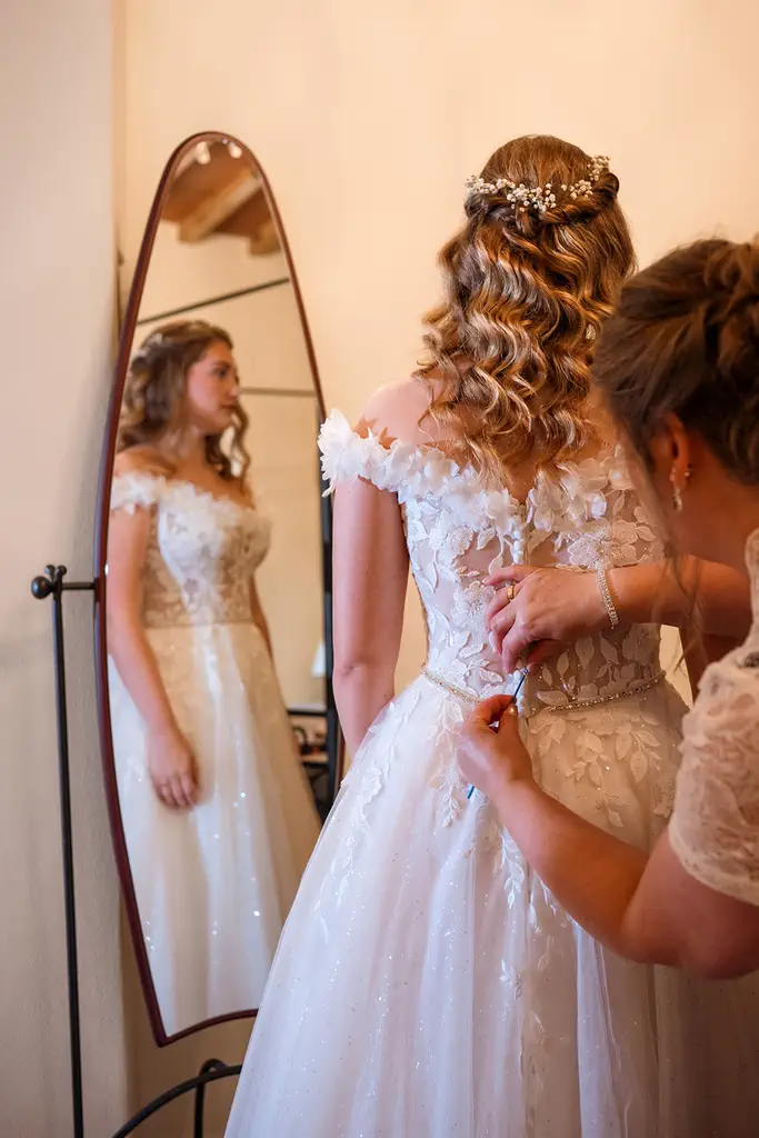 Victoria and Kevin's Monte Amiata wedding A woman in a white wedding dress has her gown fastened by another person while looking at her reflection in a mirror.